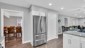 A modern kitchen with white cabinetry, a stainless steel refrigerator, wooden dining chairs, and a granite countertop. - 5 Bedroom Lavish Summer Oasis with Inground Pool (Ocean Township)