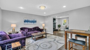 A living room with a purple sofa set, a coffee table, and a dining table with chairs. There is a fireplace, a chandelier, and a cityscape painting on the wall. - 5 Bedroom Lavish Summer Oasis with Inground Pool (Ocean Township)