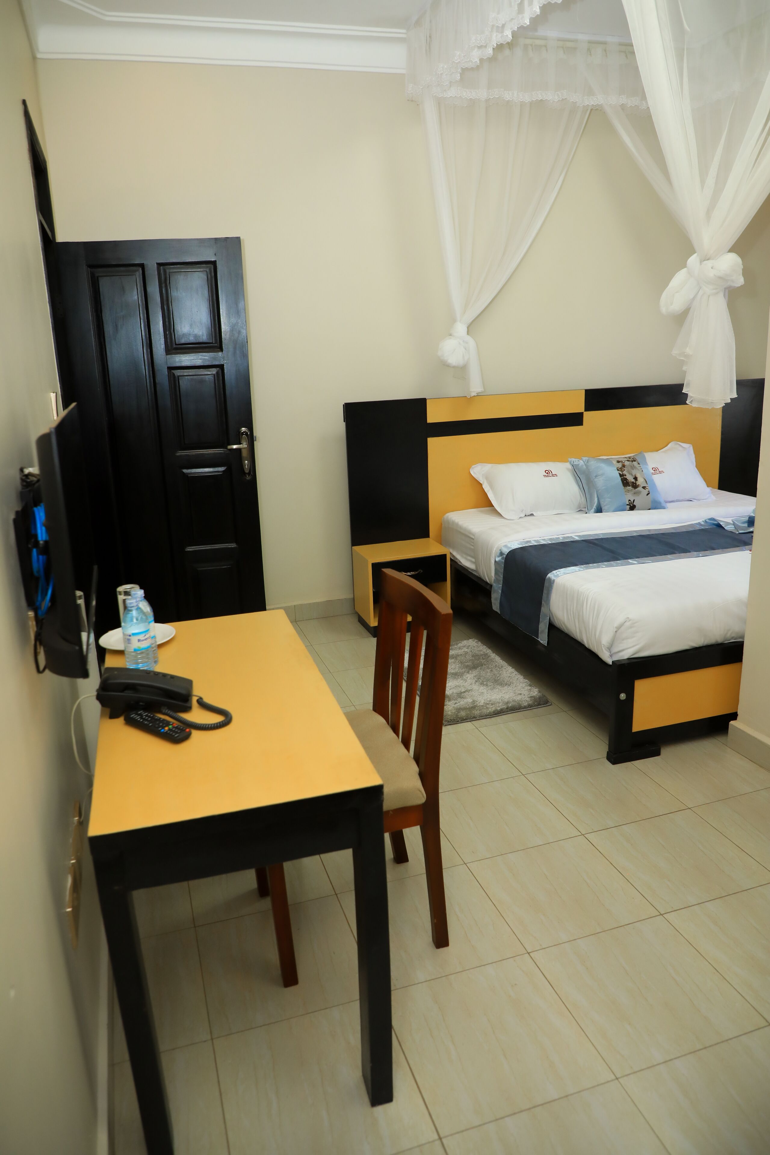 A hotel room with a bed, bedside table, chair, and a desk with a telephone and water bottle.