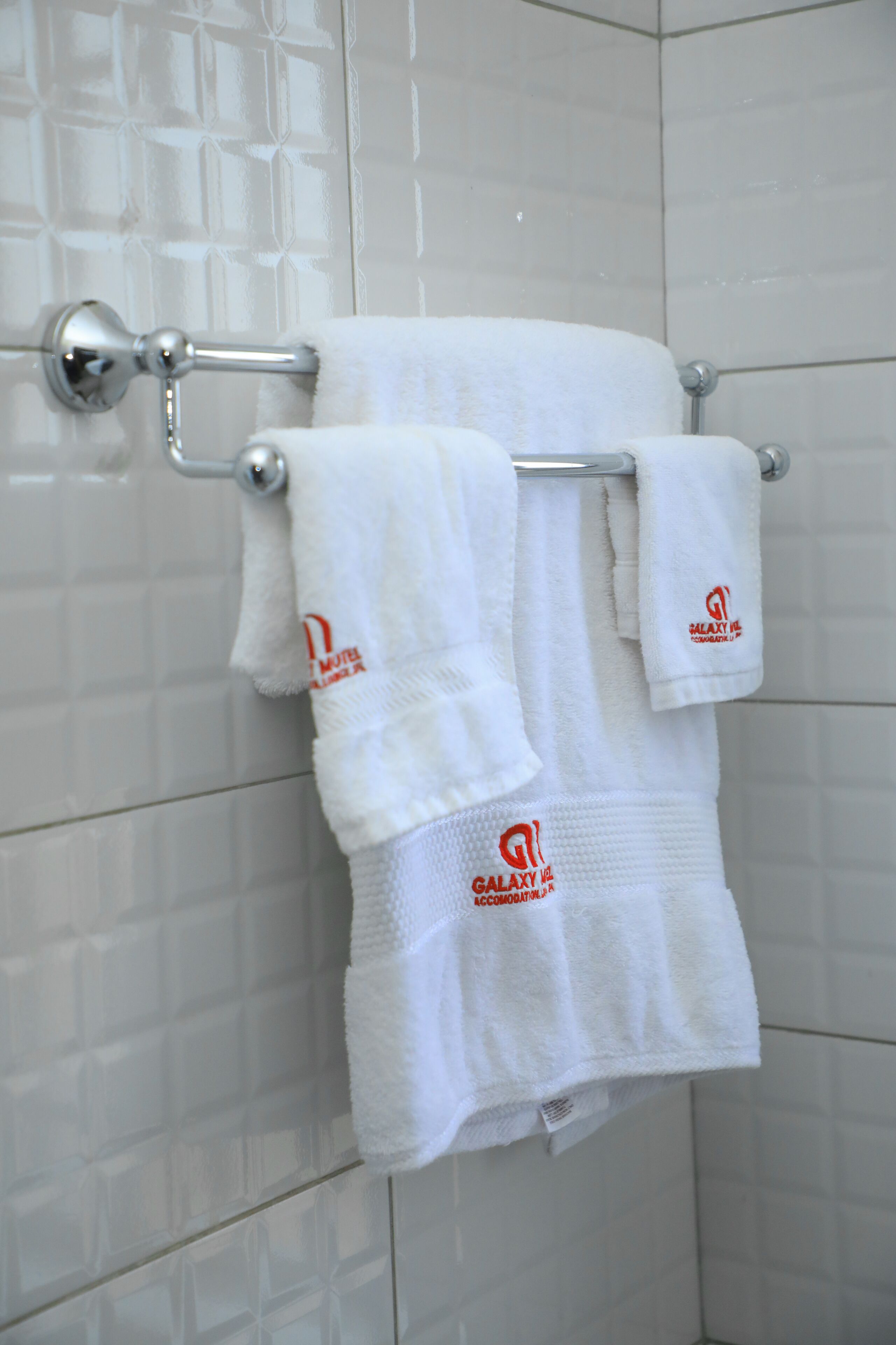 A bathroom with white tiled walls and a towel rack holding three white towels with a logo.