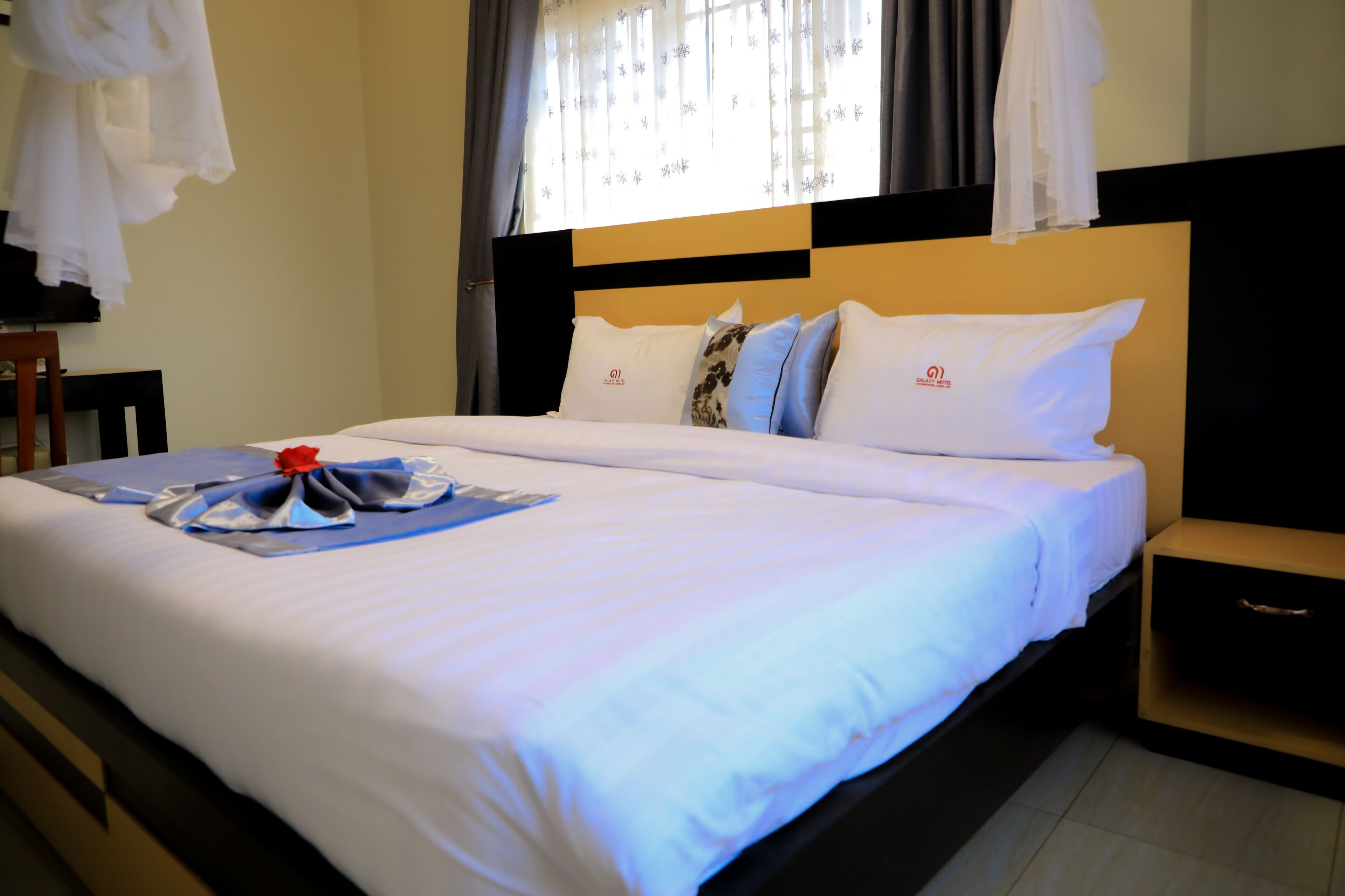 A neatly made bed with a white comforter and pillows, a bedside table, and a window with curtains.