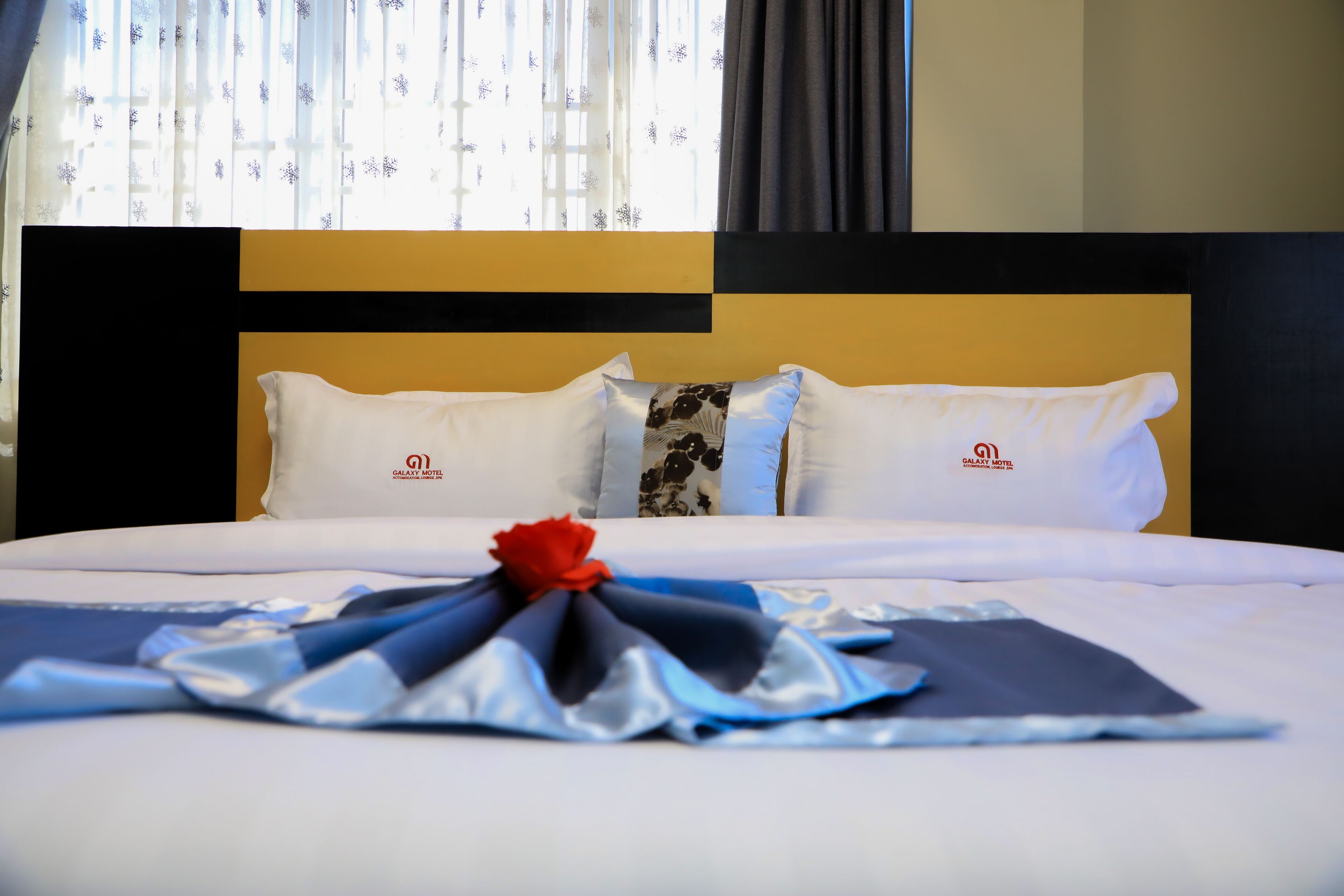 A hotel room with a neatly made bed, a decorative bow on the bedspread, and a window with curtains.