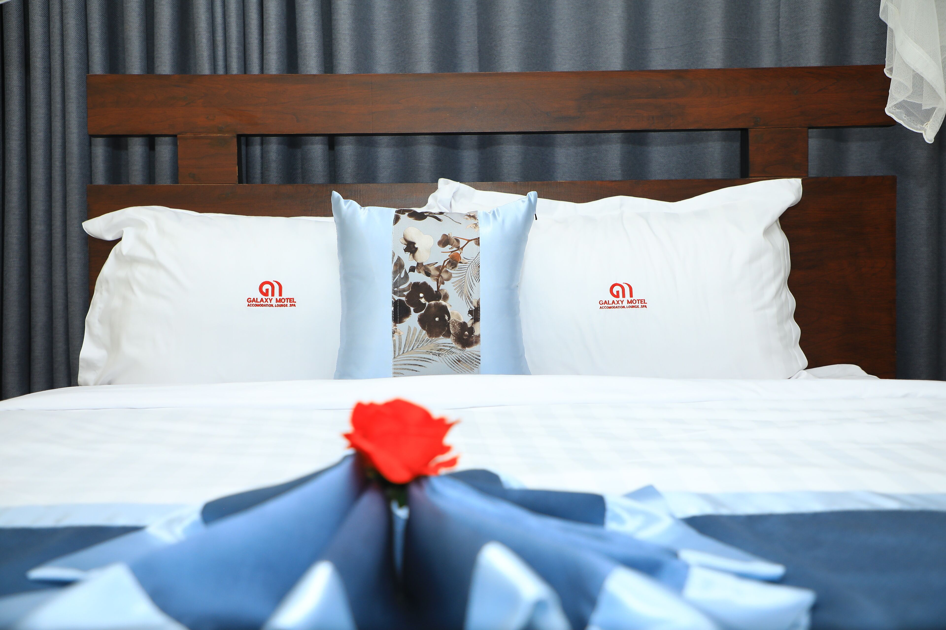 A neatly made bed with white pillows and a floral-patterned pillow, a red rose, and a blue ribbon.