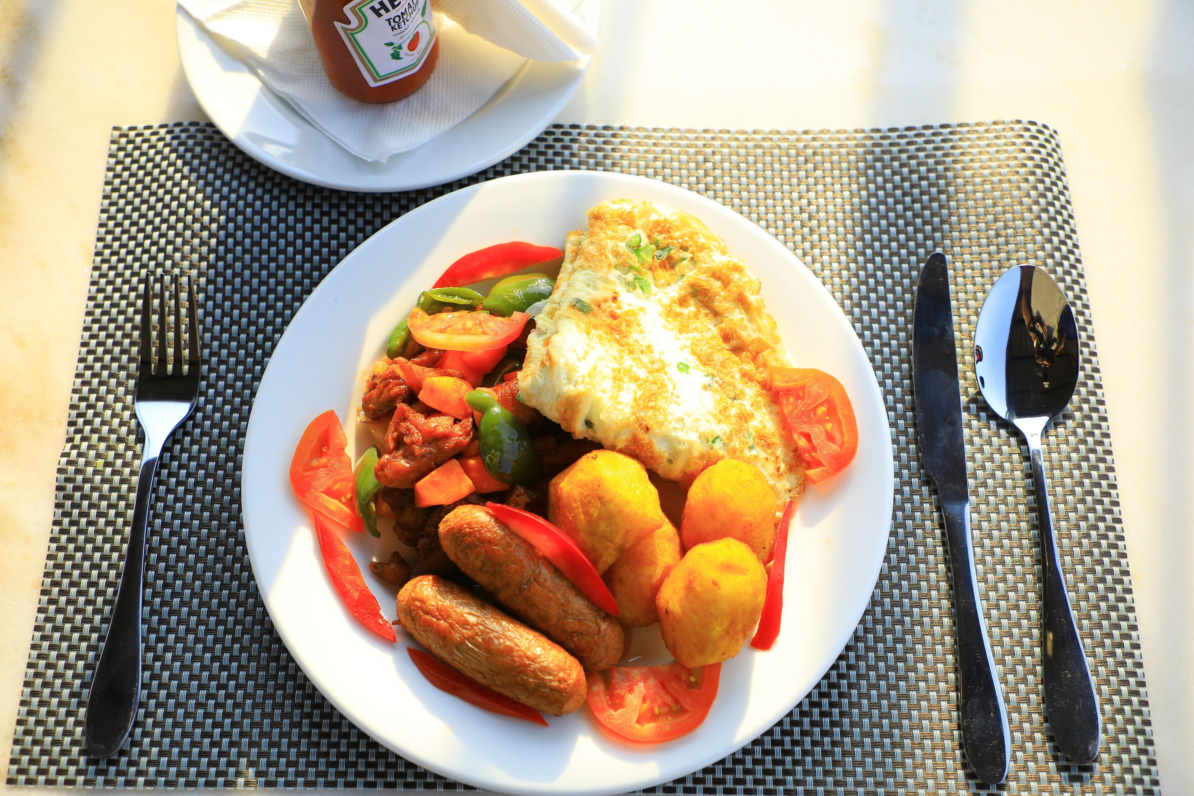 Un plato de comida con un trozo de pescado, verduras y salchichas sobre una mesa con un mantel individual.