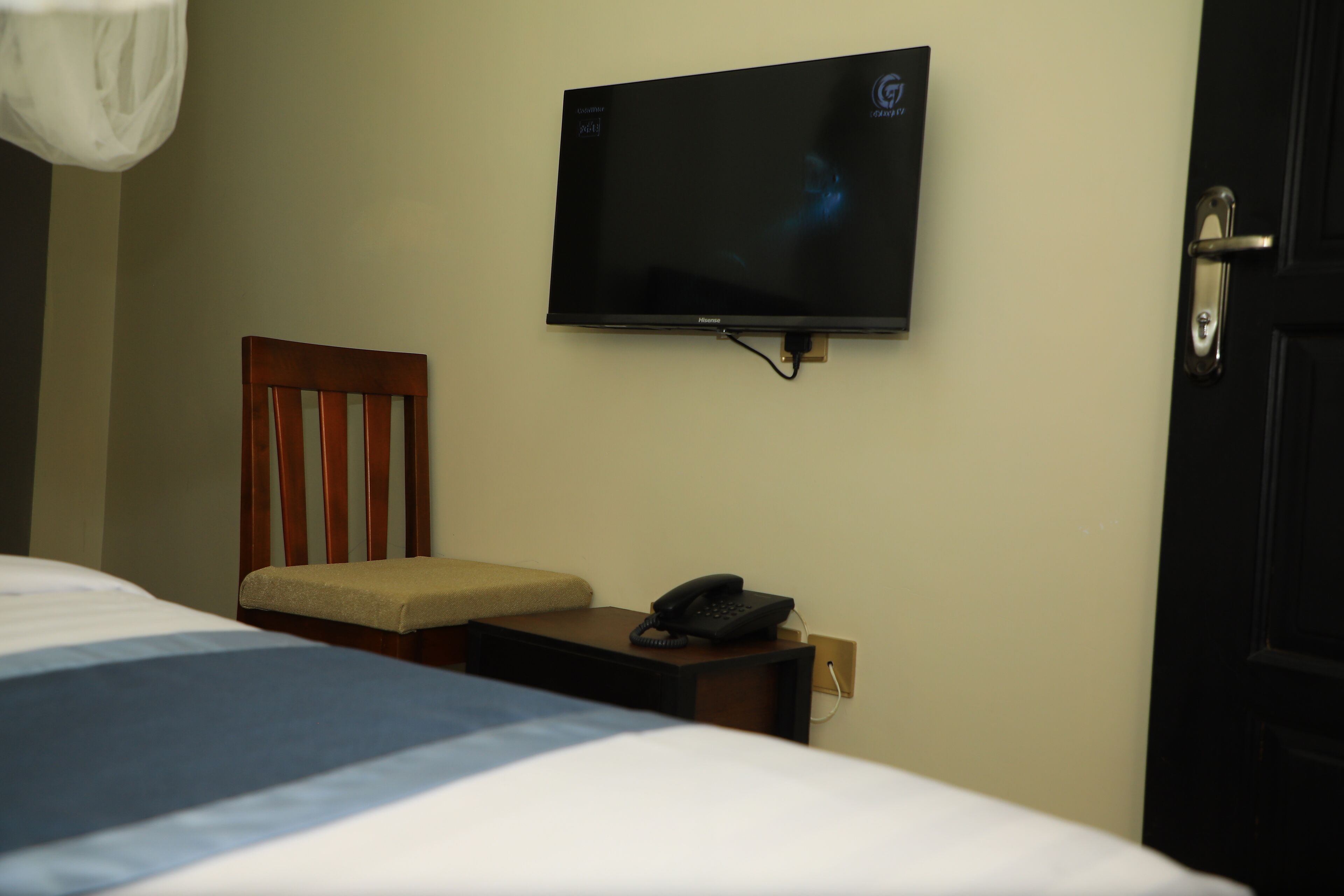Ein Hotelzimmer mit einem Bett, einem Holzstuhl, einem Nachttisch mit Telefon und einem an der Wand befestigten Fernseher.