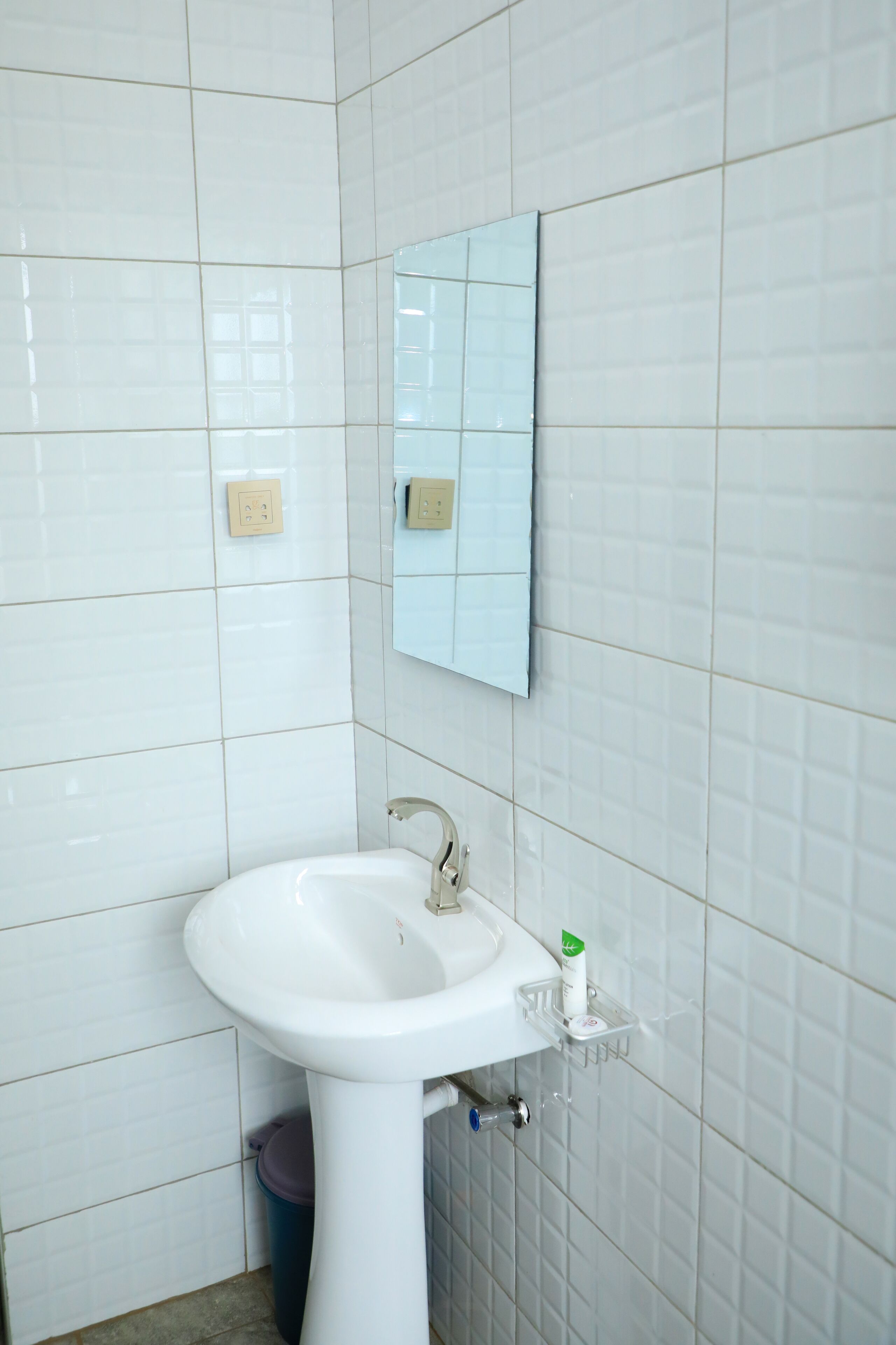 Baño con paredes de azulejos blancos, un lavamanos blanco con espejo encima y un cesto de basura debajo.