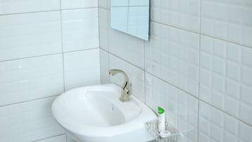 Baño con paredes de azulejos blancos, un lavamanos blanco con espejo encima y un cesto de basura debajo.