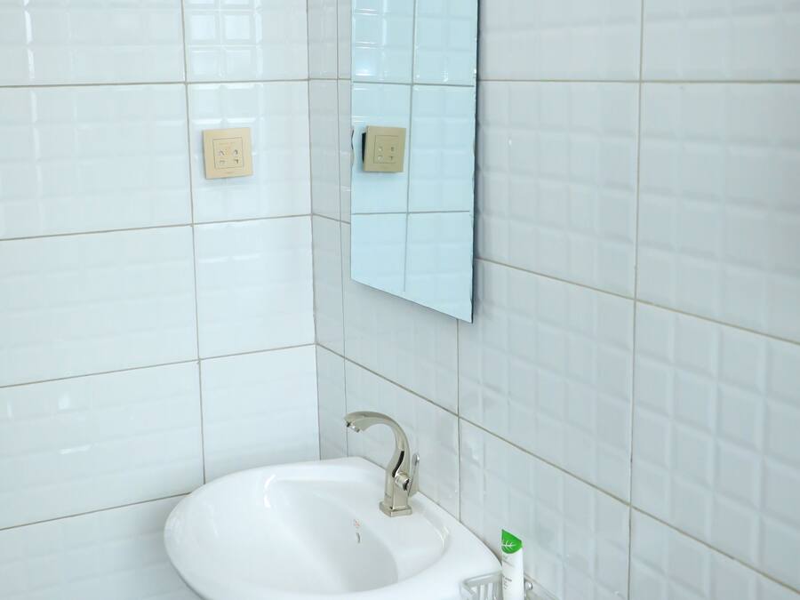 Baño con paredes de azulejos blancos, un lavamanos blanco con espejo encima y un cesto de basura debajo.