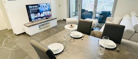 Un comedor moderno con una mesa grande dispuesta para cuatro, un televisor de pantalla plana mostrando una película y un balcón con vista a la ciudad.