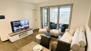 A modern living room with a flat-screen TV, a dining table set for two, and a large window offering a city view. - Luxury Flat with Parking & Large Terrace (Luxembourg City)