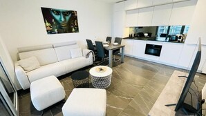 A modern living room with a sofa, ottoman, and coffee table, a dining area with chairs, and a kitchen with cabinets and a window. - Luxury Flat with Parking & Large Terrace (Luxembourg City)