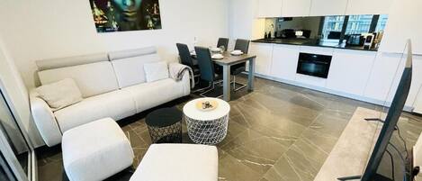 A modern living room with a sofa, ottoman, and coffee table, a dining area with chairs, and a kitchen with cabinets and a window.