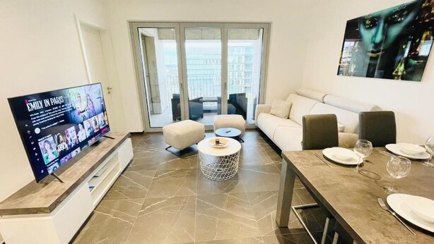 A modern living room with a flat-screen TV, a sofa, a dining table with chairs, and a balcony with a view. - Luxury Flat with Parking & Large Terrace (Luxembourg City)