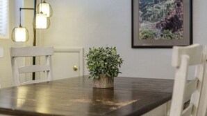 Un tavolo da pranzo con una pianta in vaso, sedie bianche e un dipinto paesaggistico incorniciato al muro.