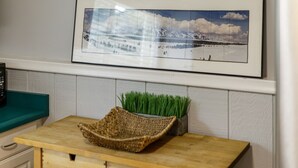 Un dipinto paesaggistico incorniciato appeso al muro, un cesto intrecciato su un tavolo di legno e una pianta verde in vaso.