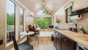 A compact, modern kitchen with a dining area, large windows, and a view of greenery.
