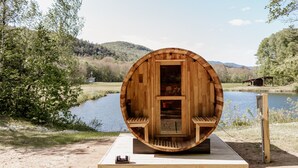 A wooden sauna structure with a bench and a door, situated by a lake with a wooden deck in front.