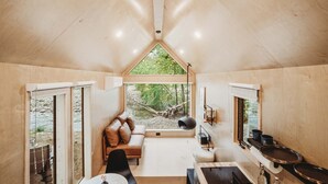 Interior acogedor de una cabaña con techo de madera, un sofá con cojines, una mesa de comedor y vistas a un bosque a través de grandes ventanales.