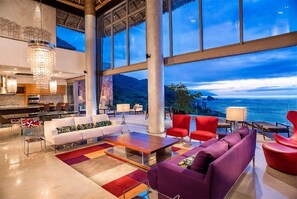 Amplia sala de estar con un ventanal que ofrece vistas panorámicas al océano, una cocina moderna y un candelabro.