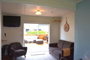 A living room with a sofa, armchair, and a television. There is a sliding glass door leading to an outdoor area with a table and chairs.