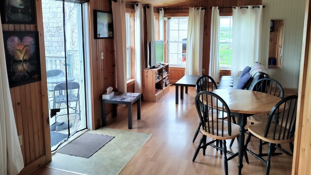 A cozy cabin interior with wooden walls and floors, a dining area with chairs and a table, a sofa, and a television on a stand. - Riverside Retreat of Mavillette (Mavillette)