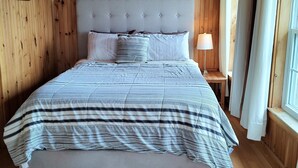 A wooden-paneled bedroom with a large bed, a bedside lamp, and a window with curtains. - Riverside Retreat of Mavillette (Mavillette)