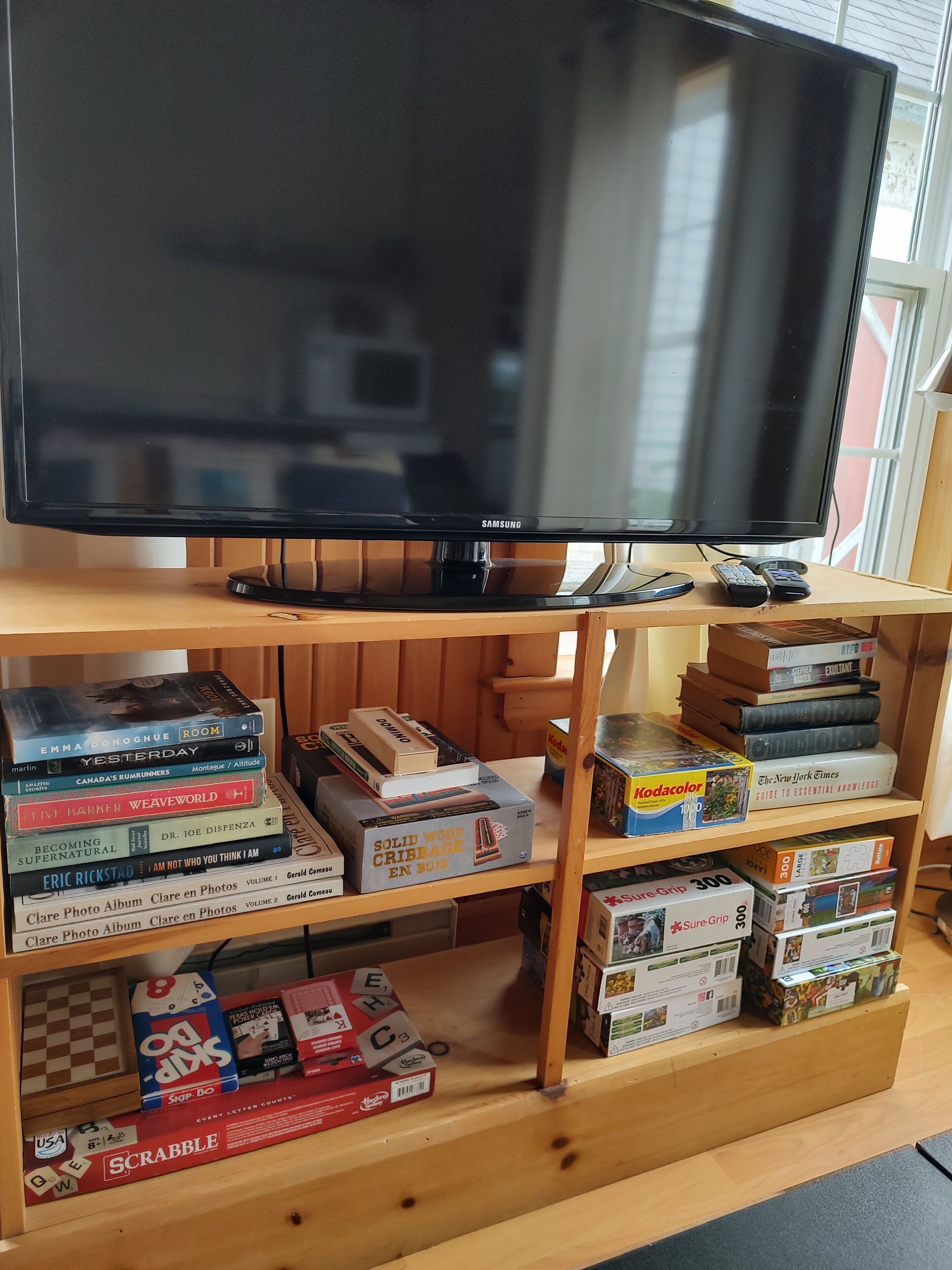 Television seisoo puisen jalustan päällä, ja sen alla on hylly, jossa on erilaisia lautapelejä ja kirjoja.