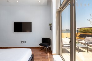 A modern hotel room with a bed, a TV mounted on the wall, a chair, and a balcony with outdoor seating.