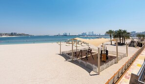 Une plage avec une aire de jeux, des palmiers et, en arrière-plan, la silhouette d’une ville.