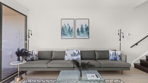 Een moderne woonkamer met een grijze modulaire bank, twee ingelijste kunstwerken, een glazen salontafel en een bijzettafeltje met een vaas.