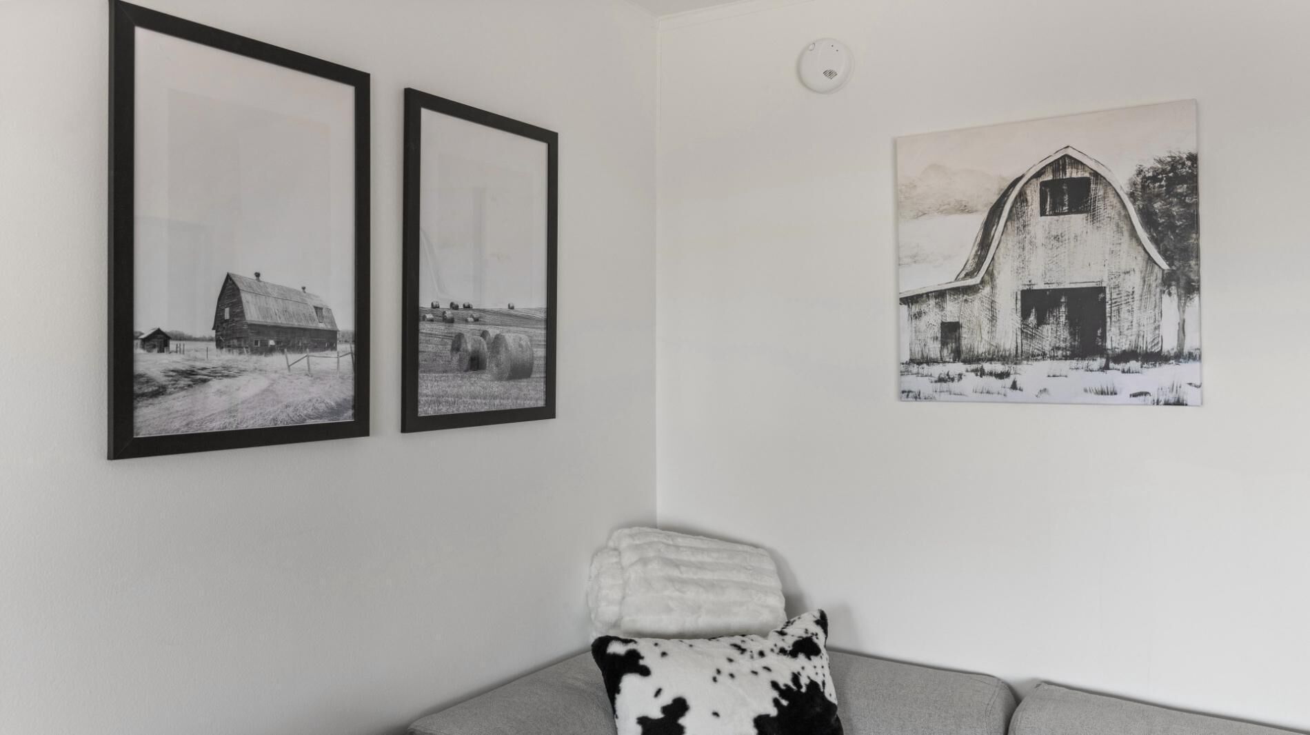 Una habitación con dos fotografías en blanco y negro enmarcadas de establos, una pared blanca y un sofá gris con un cojín estampado con motivos de vaca.