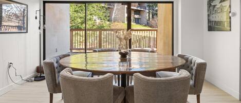 A round dining table with six chairs, a vase with flowers, and a view of a wooden deck and trees through large glass doors.