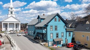 A blue building with a staircase leading to the entrance, a white building with a steeple, and a road with parked cars. - Location! This Stunning 2 Bedroom on Main Street. (Stowe)