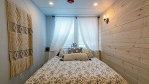A bedroom with a bed, a wall-mounted macrame piece, a window with sheer curtains, and a wooden wall. - Location! This Stunning 2 Bedroom on Main Street. (Stowe)
