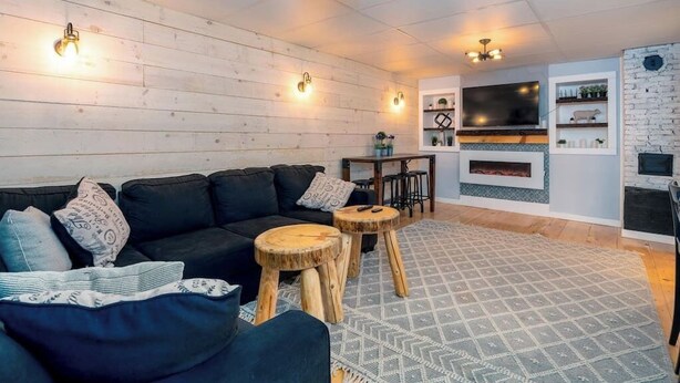 A cozy living room with a black sofa, wooden stools, a patterned rug, and a fireplace. - Location! This Stunning 2 Bedroom on Main Street. (Stowe)