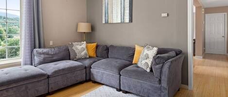 A modern living room with a grey sectional sofa, a patterned rug, and a view of greenery through a large window.