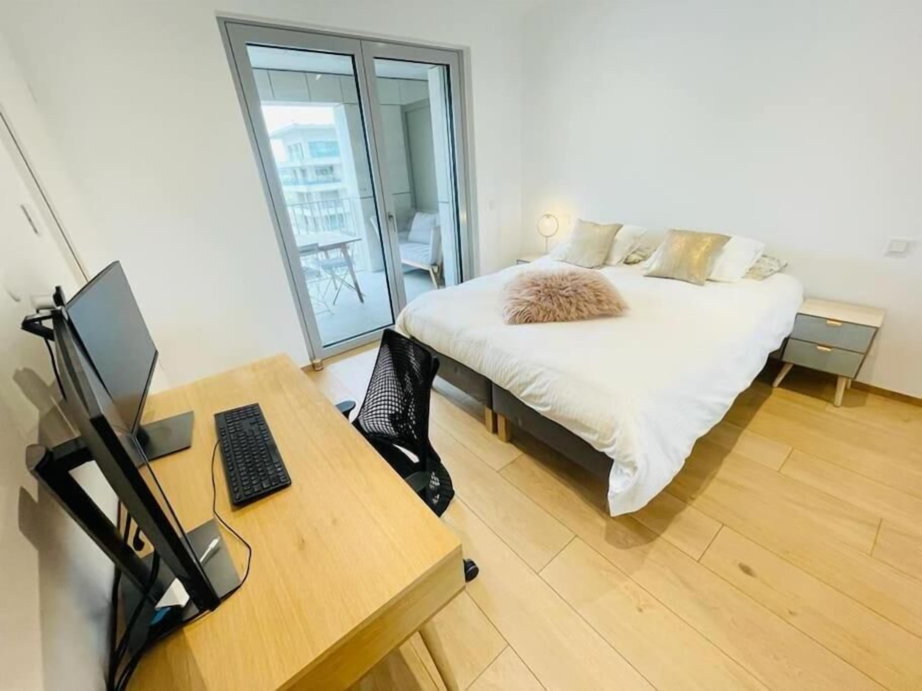 A bedroom with a bed, a desk with a computer, a chair, and a nightstand.