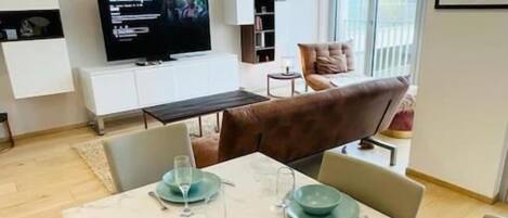 A modern living room with a flat-screen TV, a sofa, and a dining area set with green plates and glasses.