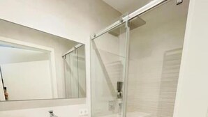 Uma casa de banho moderna com toucador branco, um espelho grande, cabine de duche em vidro e um pequeno contentor de lixo.