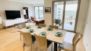 Uma área de jantar moderna com uma mesa grande posta para seis pessoas, rodeada por cadeiras. Uma televisão de tela plana está fixada na parede, e há uma varanda com vista para o exterior.
