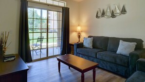 A living room with a dark blue sofa, a wooden coffee table, and a lamp. There is a window with curtains, a ceiling fan, and a view of the outdoors. - Florida Vacation Condo - No Resort Fees. Gated Resort, Gym, Pool, Hot Tub (Davenport)