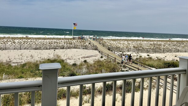 A beach with a wooden boardwalk leading to the ocean, a flag, and people walking.