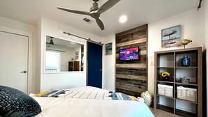A bedroom with a bed, a ceiling fan, a TV mounted on a wooden panel, and a shelf with decorative items. - Waterfront! Private, Sunrises & Sunsets, Sea life, Deck, 10 minutes to Destin! (Niceville)