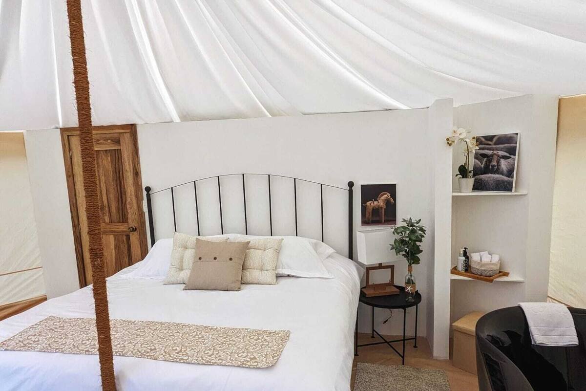 Quarto com cama, mesa de cabeceira, cadeira, prateleira pequena e porta.