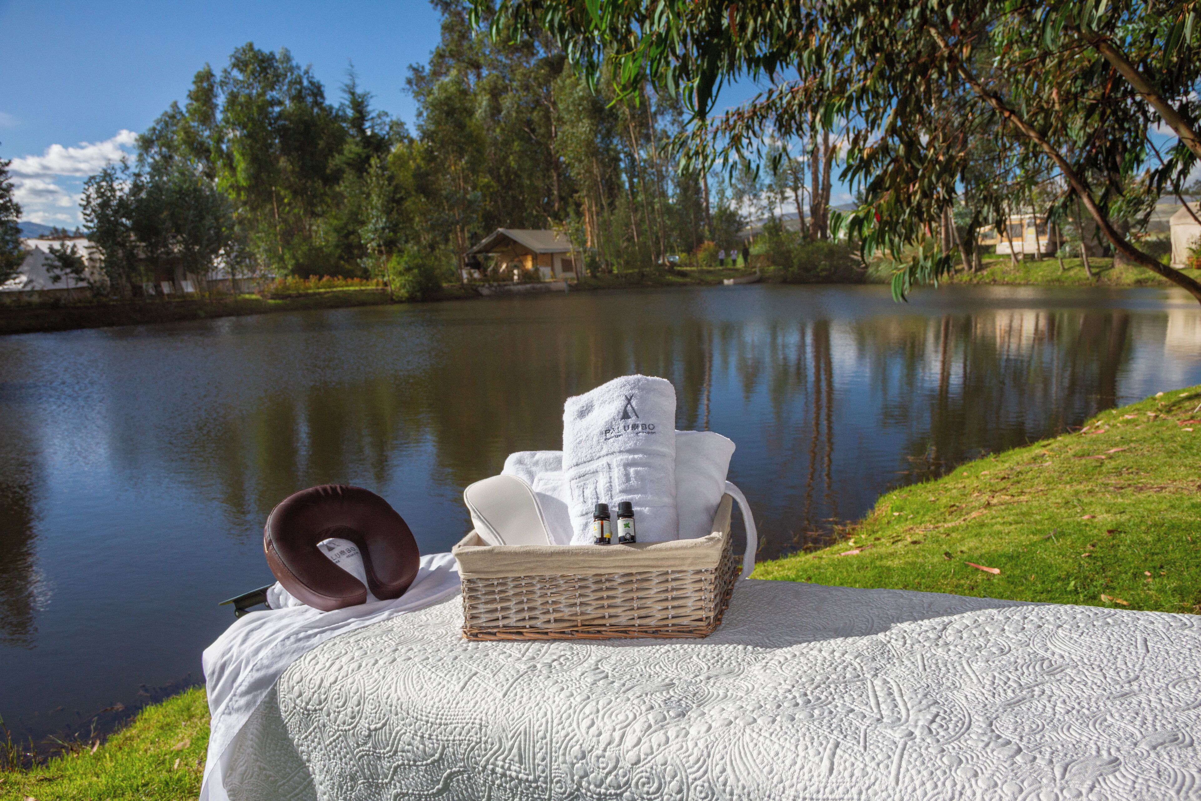A wicker basket with towels and a neck pillow on a textured white blanket by a calm lake.