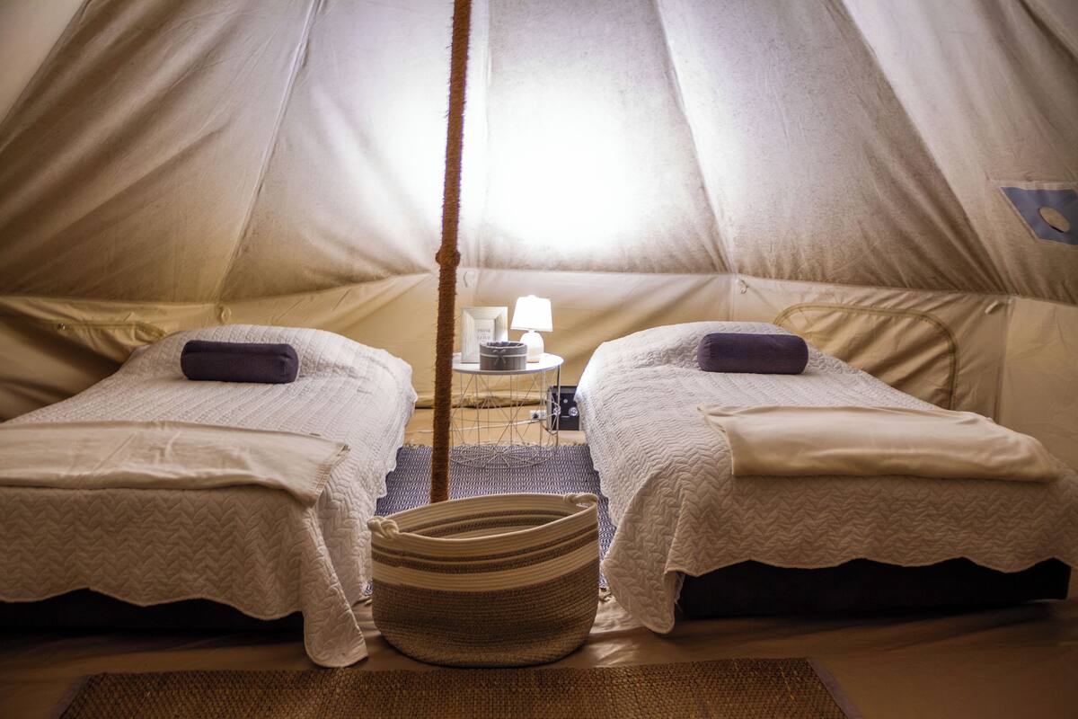 Um quarto tipo tenda com duas camas, uma mesa pequena com abajur e uma cesta de palha.