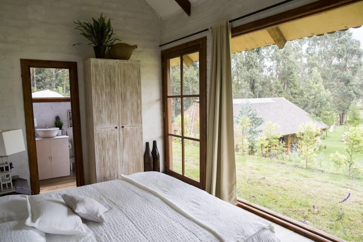 Quarto com uma cama grande, um guarda-roupa de madeira, um banheiro visível através de uma porta aberta, e vista para um jardim e uma casa lá fora.