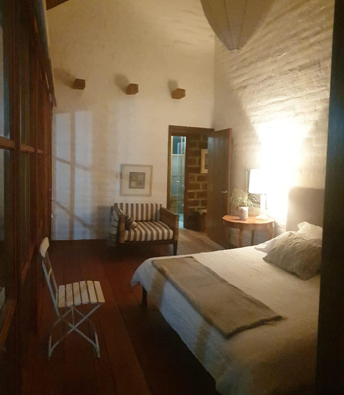 A bedroom with a bed, a chair, a small table with a lamp, and a picture on the wall.