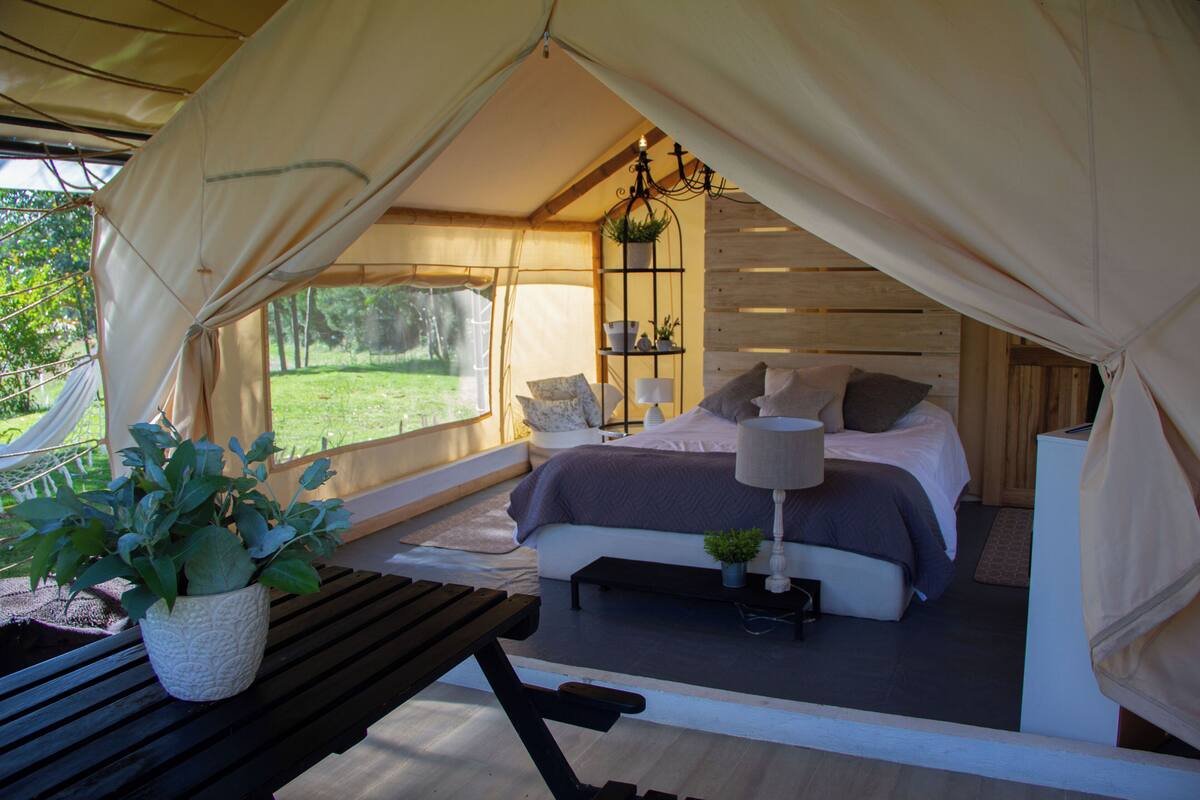 Alojamento tipo tenda com cama, abajures, uma mesinha e uma planta em vaso.