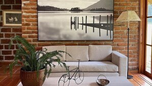 A cozy living room with a brick wall, a large framed picture of a lake, a sofa, a coffee table with a plant and a bicycle, and a floor lamp. - Palumbo Glamping & Villas (Cangahua)